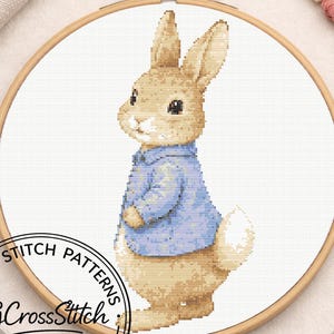 May include: A cross stitch pattern featuring a tan rabbit wearing a blue jacket, displayed within a circular wooden embroidery hoop. The design is stitched on white fabric, with the text CROSS STITCH PATTERNS CGCrossStitch visible.