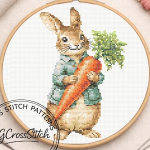 May include: Cross-stitch pattern featuring a rabbit holding a carrot. The rabbit is wearing a blue jacket and is set against a white background within a wooden embroidery hoop. The text "CG CrossStitch" is in the lower left corner.