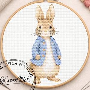 May include: A cross-stitch pattern of a rabbit wearing a blue jacket, set in a wooden embroidery hoop. The rabbit is beige and white, with black whiskers. The hoop is surrounded by embroidery floss and other craft supplies.