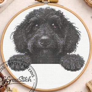 Op de afbeelding: Een kruissteekpatroon van een zwarte hond met een krullende vacht, in een houten borduurring. Het gezicht van de hond staat centraal, met zijn poten op een witte ruimte. Ambachtelijke benodigdheden omringen de ring.