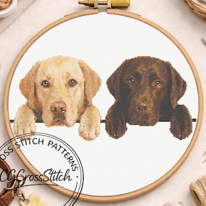 May include: A cross stitch pattern in a wooden embroidery hoop featuring two Labrador dogs, one yellow and one chocolate brown, resting their front paws on a horizontal line. The design includes the text CROSS STITCH PATTERNS CGCrossStitch.