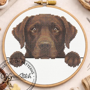 Pode incluir: Um padrão de ponto cruz de um cão Labrador Retriever castanho num bastidor de madeira. A cabeça e as patas do cão são visíveis, com um espaço branco por baixo para texto. O texto "Cross Stitch Patterns" e o nome da loja estão num círculo na parte inferior.