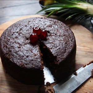 May include: A round, dark brown cake with a slice cut out, topped with red maraschino cherries. The cake is on a wooden chopping board with a silver knife.