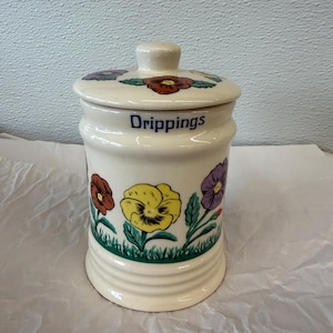 May include: Cream-colored ceramic jar with a lid, featuring a floral design of red, yellow, and purple pansies. The word "Drippings" is printed in blue on the jar. The jar is decorated with green grass at the base of the flowers.