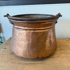 Peut inclure: Un pot en cuivre martelé avec un large bord et deux petites poignées. Le pot a un corps arrondi et une patine sombre et vieillie. Le pot est posé sur une surface en bois.