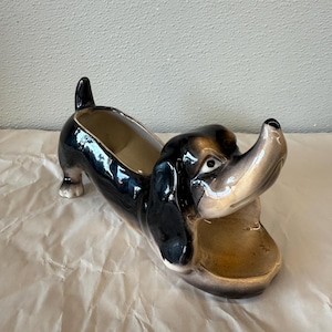 May include: A ceramic dachshund-shaped planter with a glossy black and tan finish. The dog planter has an open mouth and a hollow interior for holding plants or other items. The planter is resting on a white, textured surface.