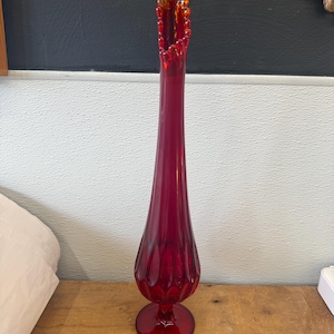 Puede incluir: Un jarrón alto de cristal rojo rubí con un borde acampanado y rizado. El jarrón tiene un cuerpo delgado y alargado con una base bulbosa y un pie redondo. El borde tiene un tono ámbar dorado. El jarrón mide unos 30 cm de alto.