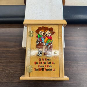 May include: Wooden box with a hinged door featuring a colorful illustration of two cartoon cowboys. The door has the text "If At Home You Do Not Find Us. Leave A Note That Will Remind Us."