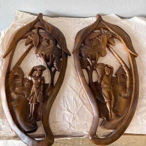 May include: A pair of wooden wall sculptures depicting tropical scenes. Each sculpture features a carved figure, palm trees, and a hut. The sculptures are brown and have a rustic, hand-crafted look.