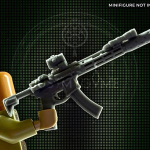 Puede incluir: Un rifle de juguete gris con mira telescópica y cargador es sostenido por una minifigura. El rifle tiene un diseño detallado con múltiples componentes. El fondo es verde oscuro con un patrón de pequeños puntos. El texto "MINIFIGURE NOT INCLUDED" está en la parte superior.