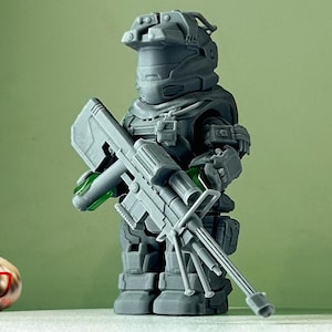 Könnte beinhalten: Eine graue Plastik-Actionfigur eines Soldaten mit Helm, der ein großes graues Gewehr hält. Die Figur steht auf einer weißen Oberfläche.
