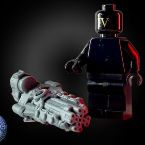 Op de afbeelding: Een grijs plastic speelgoedwapen met een zwarte handgreep en meerdere vaten. Het wapen ligt op een zwart oppervlak. Een zwart Lego-minifiguur met een gouden 'IV' op zijn borst staat achter het wapen.