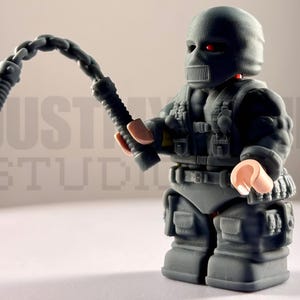 May include: A dark gray plastic toy figure wearing a tactical vest, belt, and a full-face mask with red eye lenses. The figure holds a dark gray chain whip in one hand. The figure has light-colored hands and stands in a rigid, upright position.