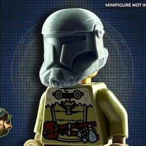 Puede incluir: Un casco gris es usado por una minifigura de Lego. La minifigura tiene un torso color beige con un diseño detallado. El fondo es azul oscuro con un patrón de cuadrícula. El texto "MINIFIGURE NOT INCLUDED" está en la parte superior.