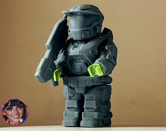 May include: A gray 3D printed action figure of a soldier wearing a helmet and holding a gun. The figure has green accents on the hands and gun.