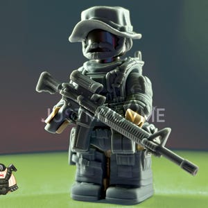 May include: A grey toy soldier figure wearing a hat, tactical vest, and holding a rifle. The figure has a serious expression and is posed against a blurred background. A small cartoon image of a vest is in the lower left corner.