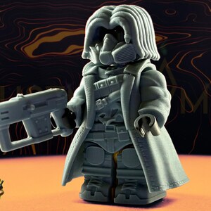 May include: A gray figurine of a character in a long coat, gas mask, and holding a futuristic gun. The figurine is standing on an orange surface with a dark background. The word "GAME" is visible in the background.