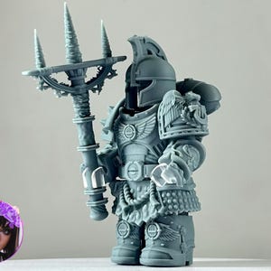 May include: A gray figurine of a warrior in detailed armor, holding a trident. The figurine has a helmet and is adorned with intricate designs, including wings and a belt. The overall theme is fantasy or science fiction.