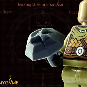 以下が含まれることがあります： グレーの岩のようなオブジェクトを持つレゴのフィギュア。フィギュアはタン色の頭と模様のある胴体を持っています。背景は暗く、「Trading With: JUSTMYGVME」と書かれています。