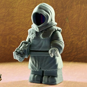 May include: A gray figurine of a hooded figure with a blocky design, reminiscent of a building block toy. The figure has a detailed cloak and holds a rectangular object. A small, cartoonish character with goggles is in the lower left corner.