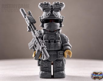 May include: A gray monochromatic toy soldier figurine featuring a tactical helmet with night vision goggles, a face mask, and a plate carrier vest. The figure holds a detailed assault rifle with a suppressor and optic sight in its right hand.