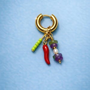 Peut inclure: Boucle d'oreille dorée avec des breloques pendantes. Les breloques comprennent un piment rouge, un bâtonnet de perles vertes et une chaîne de perles violettes, vertes et blanches. La boucle d'oreille est posée sur un fond bleu clair.