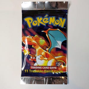 May include: Sealed Pok&eacute;mon Trading Card Game pack. The packaging is dark blue with the Pok&eacute;mon logo in yellow and orange. A Charizard illustration is featured. The pack reads "11 Tradable Game Cards."