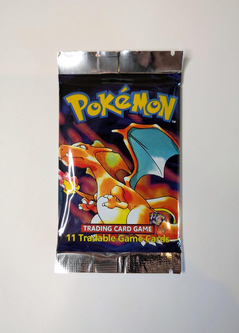 May include: Sealed Pok&eacute;mon Trading Card Game pack. The packaging is dark blue with the Pok&eacute;mon logo in yellow and orange. A Charizard illustration is featured. The pack reads "11 Tradable Game Cards."