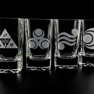 May include: Four clear shot glasses with etched designs. Each glass features a different symbol: a triangle with symbols, three connected circles, a wavy design, and a crescent moon with a circle. The glasses are arranged in a row.
