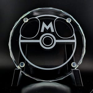 May include: A clear, faceted glass award with a circular design. The design features the letter "M" in the center, surrounded by a geometric pattern. The award is displayed on a black stand against a dark background.
