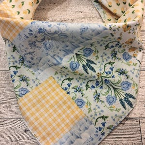 Peut inclure: Un bandana triangulaire pour animal avec un motif patchwork composé de vichy jaune, de petites fleurs jaunes et de motifs botaniques bleus avec des illustrations florales complexes sur fond blanc, noué en haut par un nœud en tissu.