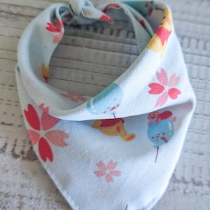 Puede incluir: Bandana azul claro con un patrón repetido de flores rosas, detalles amarillos y personajes de dibujos animados. La bandana está anudada en la parte superior y está hecha de una tela suave y ligera.