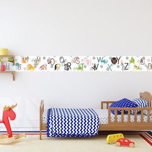 Op de afbeelding: Een witte muur met een kleurrijke alfabetrand met dieren en stippen. De rand bevindt zich boven een houten peuterbed met blauw-witte chevron beddengoed. Een houten schommelpaard bevindt zich op de voorgrond.