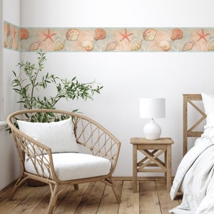May include: A white wicker chair with a white cushion sits in a room with a white wall and a wooden floor. A wooden side table with a white lamp sits next to the chair. A bed with a white headboard and white bedding is in the background. A seashell border wallpaper is on the wall.