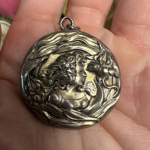 May include: A silver locket pendant with an ornate design featuring a woman's face and floral motifs.