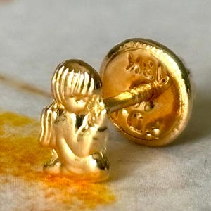 Pu&ograve; includere: Un orecchino dorato con una piccola e dettagliata figura di una persona seduta. Il perno e il retro dell'orecchino sono anch'essi dorati, con segni visibili sul retro. Il design &egrave; intricato e delicato.