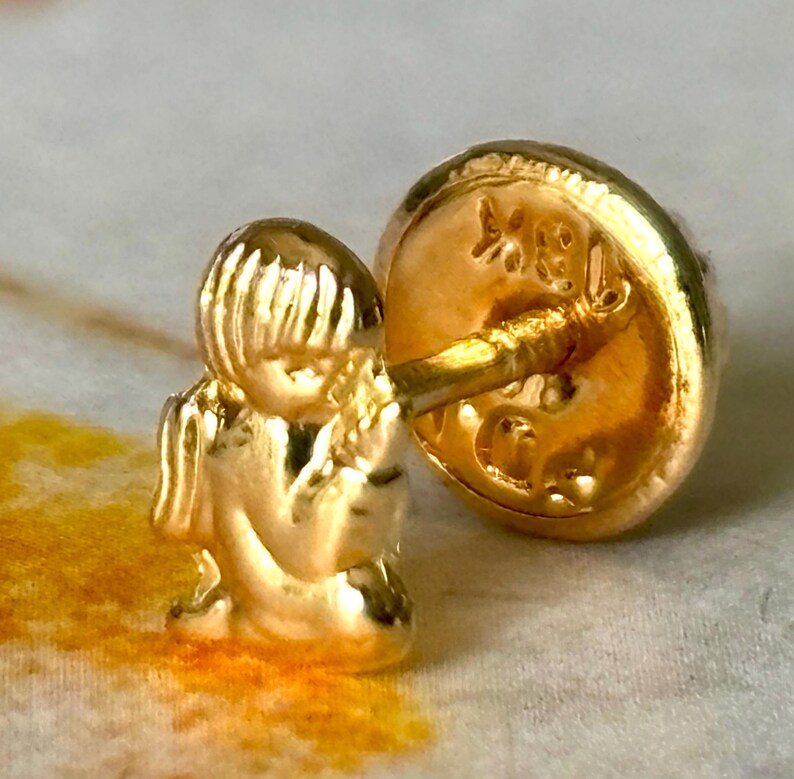 Pu&ograve; includere: Un orecchino dorato con una piccola e dettagliata figura di una persona seduta. Il perno e il retro dell'orecchino sono anch'essi dorati, con segni visibili sul retro. Il design &egrave; intricato e delicato.