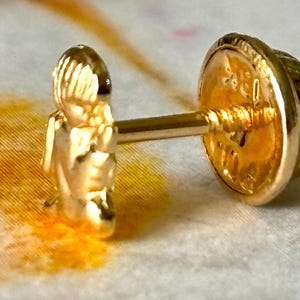 Pu&ograve; includere: Un orecchino dorato con un design dettagliato. L'orecchino presenta una piccola figura sul davanti e un retro decorativo. Il perno &egrave; visibile, su uno sfondo strutturato. L'orecchino &egrave; in oro.