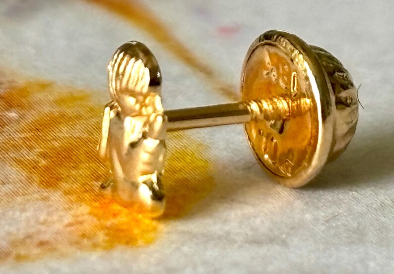 Pu&ograve; includere: Un orecchino dorato con un design dettagliato. L'orecchino presenta una piccola figura sul davanti e un retro decorativo. Il perno &egrave; visibile, su uno sfondo strutturato. L'orecchino &egrave; in oro.
