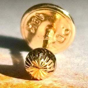 May include: Close-up of a gold earring with a textured, spherical stud and a round backing. The earring has markings on the backing, and the stud features a radial pattern. The jewelry is set against a soft, neutral background.