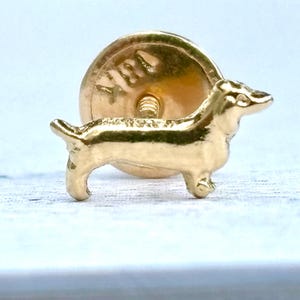 Puede incluir: Un broche de perro salchicha dorado. El broche presenta una figura detallada en miniatura de un perro salchicha, con un respaldo redondo y un cierre de alfiler. El perro está de pie, con las patas visibles. El broche es probablemente de metal.