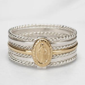May include: A stackable ring set featuring a silver band with a twisted rope design, a gold band, and a silver band with a gold oval centerpiece. The gold centerpiece has a religious figure engraved on it.