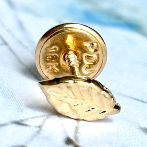 May include: Close-up of a gold-colored leaf-shaped earring stud. The earring has a detailed leaf design and a post with a round backing. The backing is stamped with "18K" and other markings. The earring is set against a soft, patterned background.