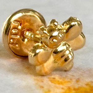 May include: Close-up of a gold-colored bee-shaped lapel pin. The pin has a round backing with a post and clasp. The bee has detailed wings and body, and the pin is on a white and yellow surface.