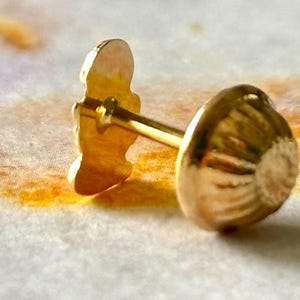 Pu&ograve; includere: Un orecchino dorato con un perno e un elemento decorativo. L'orecchino presenta una piccola figura stilizzata e un retro arrotondato e strutturato. L'orecchino &egrave; posto su uno sfondo morbido e colorato.