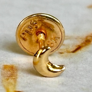 Könnte beinhalten: Ein goldfarbener Ohrstecker mit einem Halbmond-Design. Der Ohrring hat einen Stecker und eine runde Rückseite. Der Ohrring ist wahrscheinlich aus Gold oder vergoldetem Metall gefertigt. Der Ohrring wird vor einem weißen und braunen Hintergrund gezeigt.