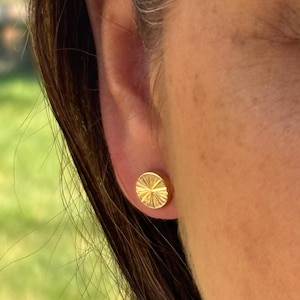May include: A close-up of a gold stud earring with a sunburst design. The earring is round and has a textured surface. The earring is on a person's ear.