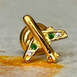 May include: A gold-colored airplane pin with green and white gemstones. The pin features a classic airplane design with a round backing. The gemstones are set into the wings and body of the plane, adding a touch of sparkle.