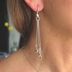 May include: Silver dangle earrings with three strands, each adorned with small gold and silver beads. The earrings are suspended from a silver hook and are shown against a blurred background.
