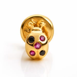 Peut inclure: Une boucle d'oreille à clou en métal doré en forme de coccinelle, dotée d'une finition polie. La surface arrondie est incrustée de trois petites pierres précieuses roses circulaires et de deux pierres noires circulaires disposées symétriquement.